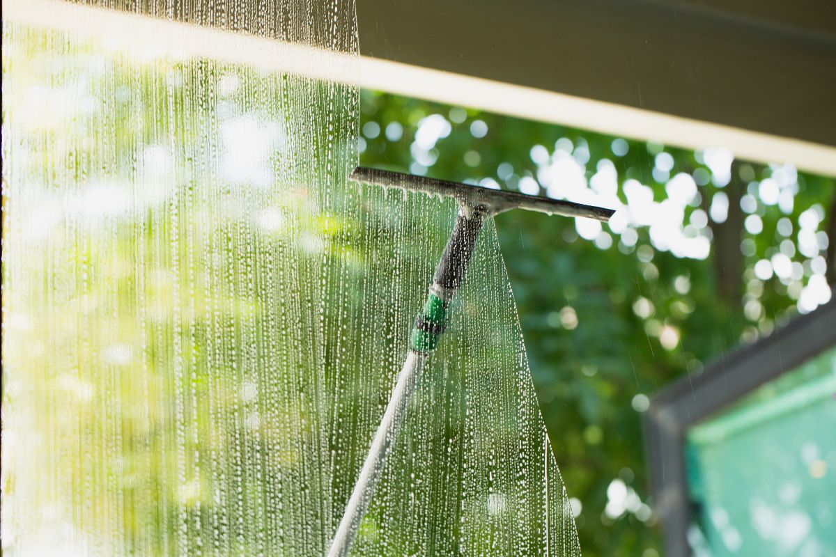 A window with a hose attached, alongside a bottle of window cleaner, set against a bright outdoor background.